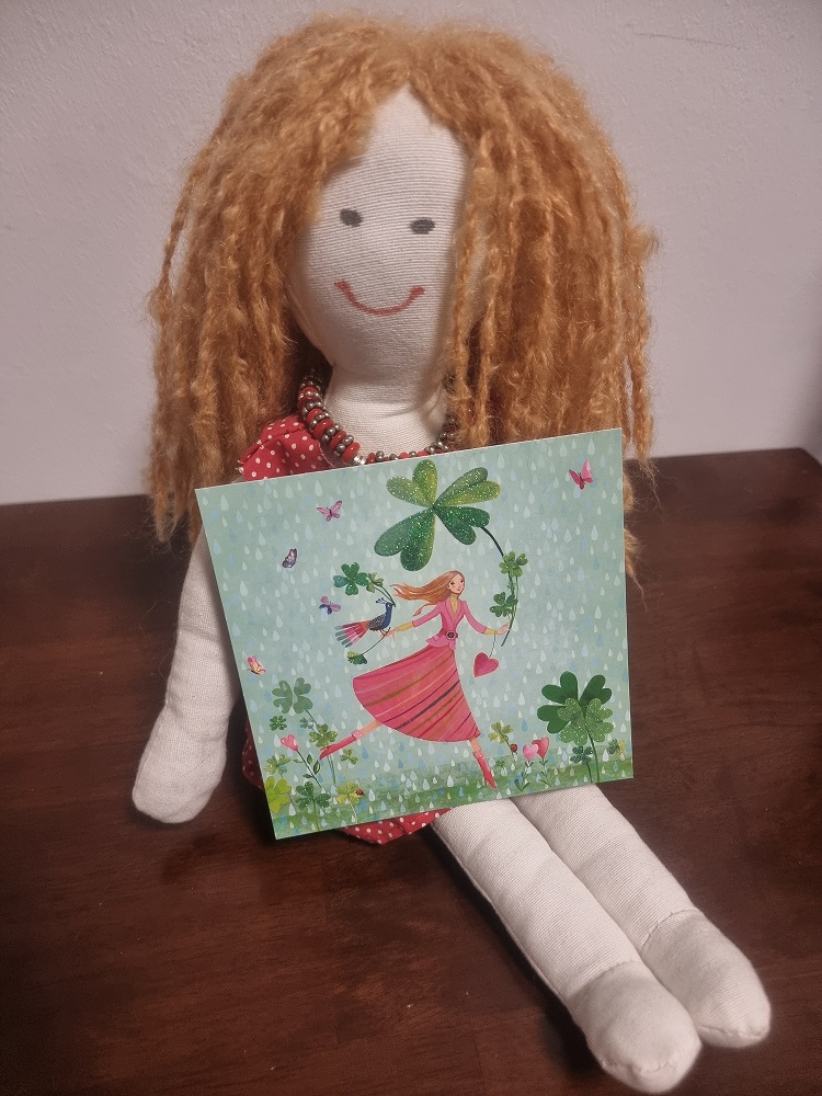 Unser Maskottchen Sophie zeigt und eine quadratische Postkarte von Mila Marquis - eine Fee, die mit Glücksklee durch die Karte läuft